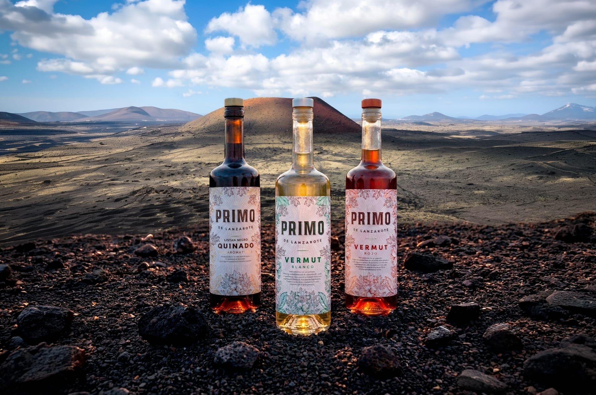 Las tres botellas de Primo de Lanzarote — Quinado, Vermut Blanco y Vermut Rojo — sobre el picón negro de Lanzarote, con conos volcánicos al fondo